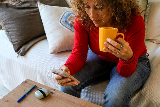 Serious mature woman with curly hair holding smartphone and coffee checking passwords at home. Adult managing digital security, accessing apps and protecting personal data in relaxed indoor lifestyle