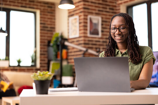 African american woman smiling brightly at device screen, reading online articles in brick wall apartment. Portrait of black female remote worker happily using laptop at desk, checking office emails.