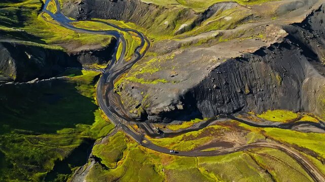 Drone panorama of a tiny car driving a winding highland road through Iceland&rsquo;s green summer valley and dark volcanic cliffs. Remote living, freedom, and adventure travel concept.