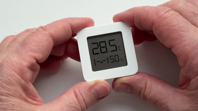 Close-up of woman's hands holding digital sensor displaying temperature and humidity, careful hands tracing display, wrinkled skin.
