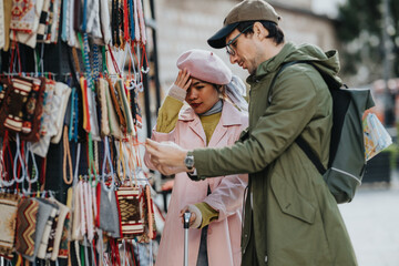Fototapeta premium Diverse tourists browse handmade bags and accessories at an outdoor street market while consulting a map. A couple examines souvenirs together, comparing options and planning their next stop.