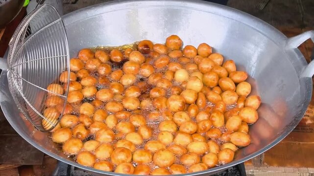 Popular Thai street food dessert "Khanom Khai Nok Krata" or small fried sweet potato balls being deep-fried in hot oil in a large wok at a local market. Thai fried sweet potato balls