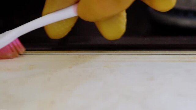 Closeup yellow grime along sink seam, gloved hand applies cotton swab to lift film, solvent action begins to break down buildup, focused domestic maintenance scene by sink rim.