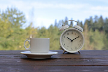 A white cup of coffee stands next to a retro alarm clock on a wooden desk against the backdrop of the spring forest. © vubaz