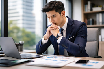 Obraz premium Young businessman in navy suit working on laptop in modern office, focused on financial documents and charts, thoughtful expression showing concentration and determination