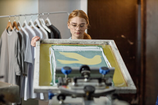 Caucasian young adult woman screen printing bird design onto fabric, standing in workshop with clothing rack in background, concentrating on aligning frame for precise application