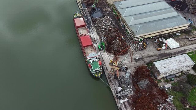 A cargo ship is at the dock in an industrial area. Workers manage the unloading of materials. Surrounding the dock are warehouses and piles of scrap materials near the water.