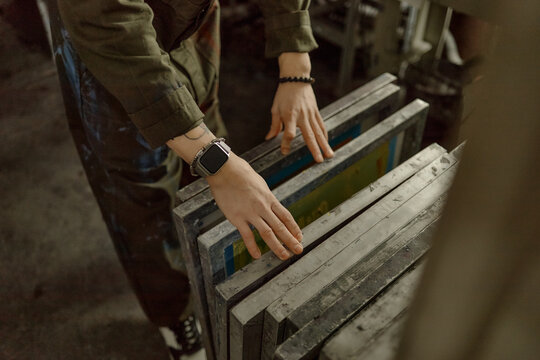 Young adult Caucasian man arranging silkscreen frames in workshop, hands visible with smartwatch and bracelet, focusing on preparing equipment for screen printing process