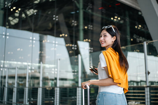Young asian woman traveler using smartphone for checking flight information check the routes trip while waiting with her luggage at the airport terminal. Traveller woman passport and baggage luggage