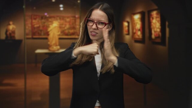Young hispanic woman with red glasses makes timeout hand gesture in an art gallery building, scowling and leaning slightly forward; art critique defiance.