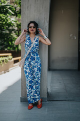 Fototapeta premium A stylish woman wears a blue floral jumpsuit with sunglasses and a cross body bag. She stands near a concrete pillar in an outdoor urban area.