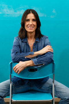 Smiling woman in blue denim shirt sitting on a chair against a blue background. Milan, Italy