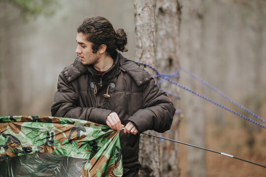 A young man adjusts a camouflage tarp while preparing a campsite in a mountain forest. He secures the tarp to trees with ropes and poles for outdoor camping.
