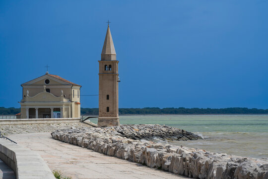 Coastal church with a tall bell tower beside a stone walkway under a clear blue sky. Caorle, Metropolitan City of Venice, Veneto, Italy