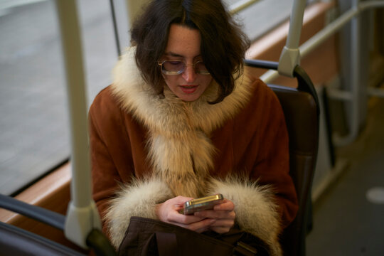 Woman in fur-lined coat using a smartphone on public transport, smiling and looking down. Brussels, Belgium