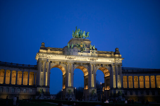 Illuminated arch monument at night against a deep blue sky with surrounding colonnades. Brussels, Belgium