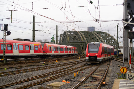 Red commuter trains on tracks with bridge and cloudy sky in the background.   Cologne, Germany