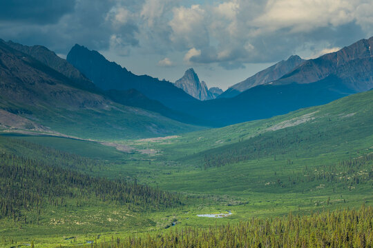 Vast green valley beneath rugged mountains under a dramatic cloudy sky. Yukon Territory, Canada