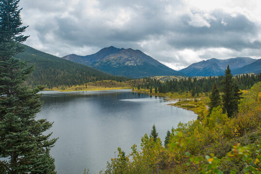 Serene mountain lake surrounded by lush trees with towering peaks under a cloudy sky. Yukon Territory, Canada