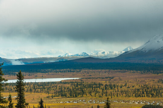 Snow-capped mountains under cloudy skies with a forested valley and serene lake view. Eureka, Alaska, USA