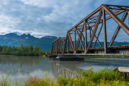 Rusted steel bridge over a calm river with mountains and trees in the background. Palmer, Alaska, USA