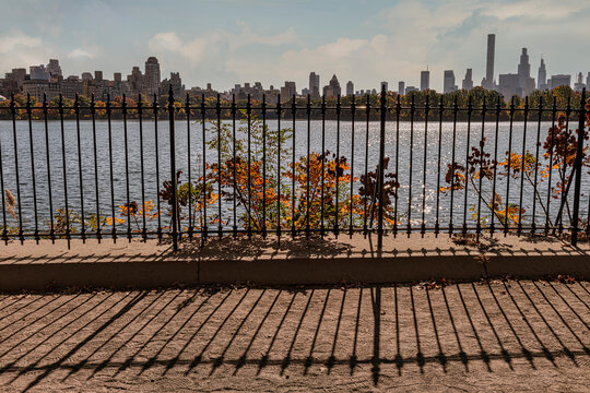 City skyline view over a serene lake, framed by a black iron fence and autumn foliage. Jacqueline Kennedy Onassis Reservoir, New York City, USA