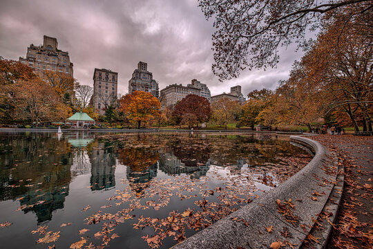 Autumn park scene with colorful trees and fallen leaves by a reflective pond and tall buildings. The Pond, Central Park, New York City, USA