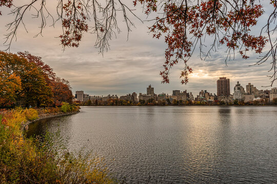 Autumn view of a city skyline across a calm lake under a cloudy sky with colorful foliage. Central Park Lake, New York City, USA
