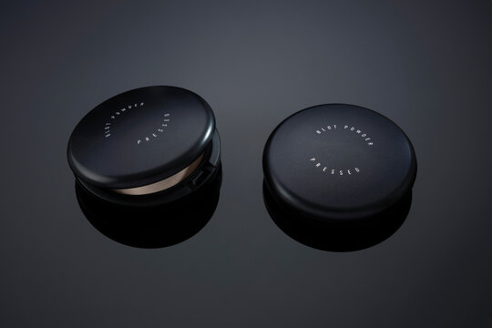 Two sleek black pressed powder compacts on a reflective dark surface.