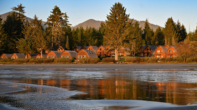 Rustic cabins with curved roofs amidst tall evergreen trees by a reflective lake at sunset. Tofino, Vancouver Island, Canada