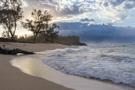 Tranquil beach with gentle waves, sandy shore, and trees under a dramatic cloudy sky. Little Paia Beach, Maui, Hawai, USA