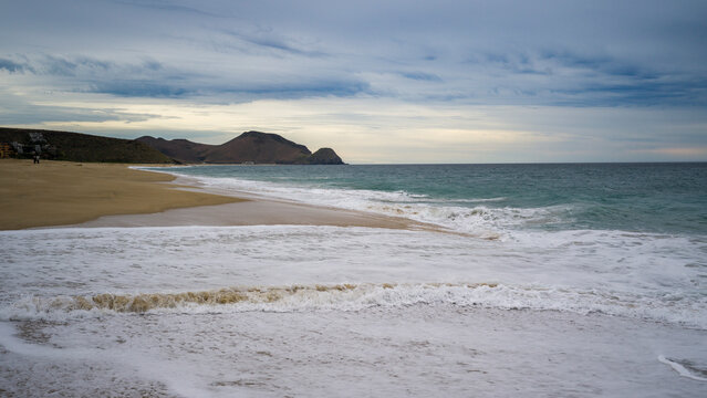 Serene beach with gentle waves, distant mountains, and a cloudy sky at dusk. Punta Labos Beach, Todos Santo, Mexico