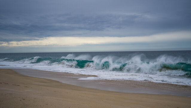 Waves crash against a sandy beach under a cloudy sky with a vast horizon in view. Todos Santos, Mexico