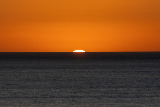 Orange sun setting over calm ocean horizon with clear sky. Todos Santos, Baja California, Mexico