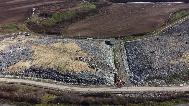 This landfill site shows large piles of waste and a road running through it. The landscape is mostly flat, with various materials scattered across the area. Some machines and workers are present.