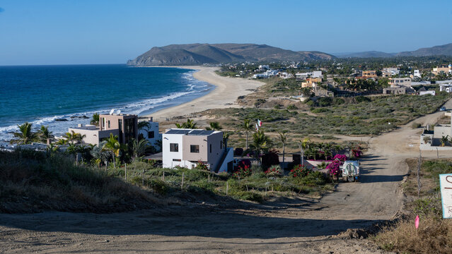 Coastal village with sandy beach, ocean waves, houses, and distant hills under a clear blue sky. Todos Santos, Baja California Sur, Mexico