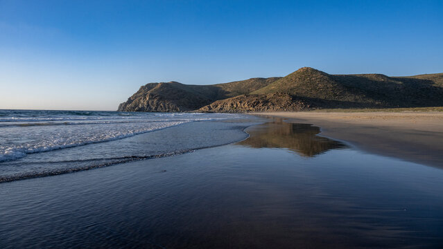 A serene beach with waves lapping against the shore under a clear blue sky and distant hills. Todos Santos, Baja California, Mexico