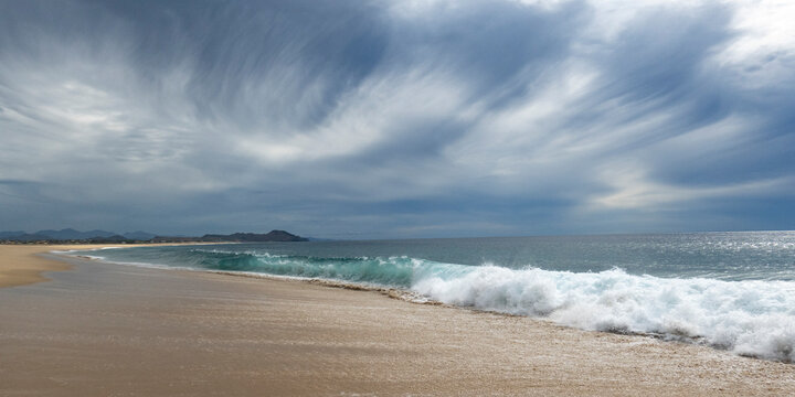 Waves crashing on a sandy beach under a dramatic cloudy sky with distant hills. Todos Santos, Baja California, Mexico
