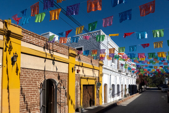 Colorful flags hang over a vibrant street with bright yellow and white buildings.   Todos Santos, Mexico