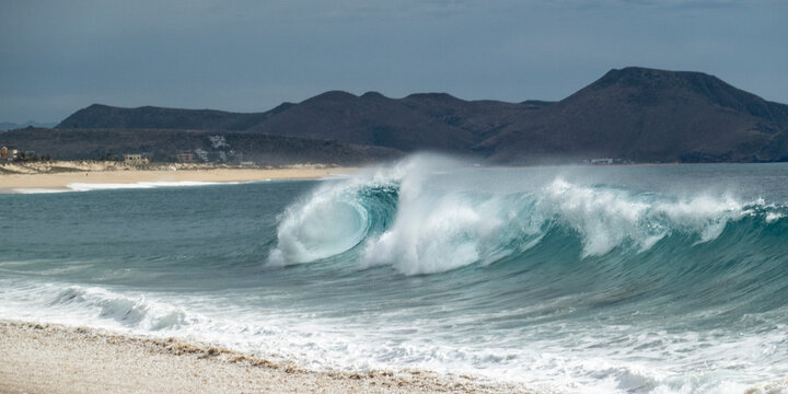 Powerful ocean waves crashing on a sandy beach with distant rocky mountains under a cloudy sky. Todos Santos, Baja California, Mexico