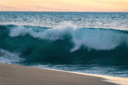 Ocean wave crashes onto a sandy beach beneath a colorful evening sky. Playa La Pastora, Todos Santos, Mexico