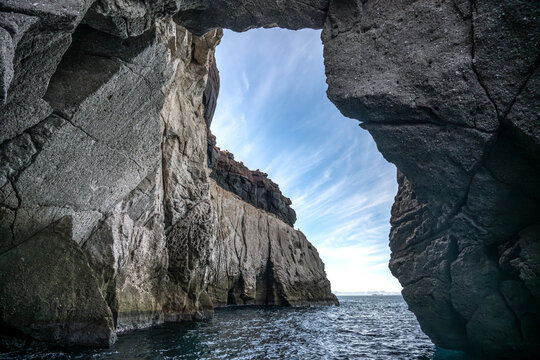 A rocky cave opening frames a view of the ocean and sky with wispy clouds above. Canada
