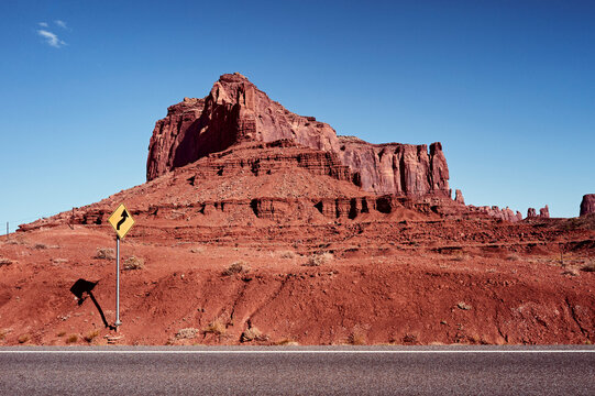 Red rock formation under a clear blue sky with a road and a caution sign in the foreground. Monument Valley, Arizona, USA