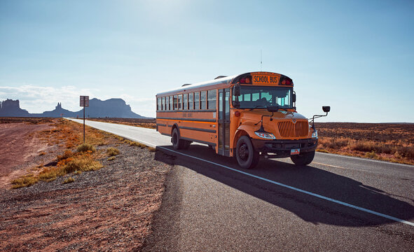 A yellow school bus drives on a deserted road through a vast desert landscape. Monument Valley, Arizona, USA