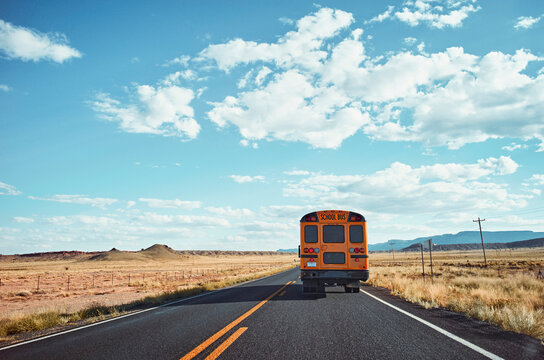 Yellow school bus travels on a long, deserted road under a bright, blue sky with clouds. Monument Valley, Arizona, USA