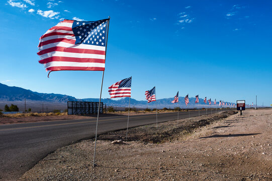American flags line a desert highway under a bright blue sky with mountains in the background. US I-15, Nevada, USA