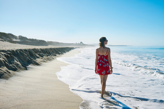 Woman in a red dress walks along a sandy beach with gentle waves under a clear blue sky. Half Moon Bay, California, USA