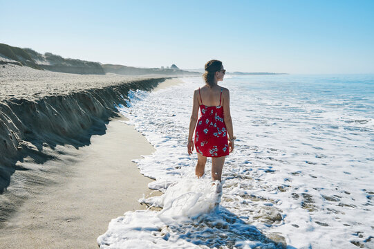 Woman in red dress walks along the beach, waves touching her feet under clear blue sky. Half Moon Bay, California, USA