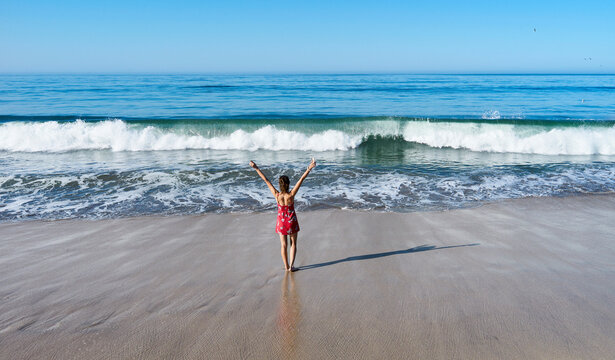 Woman in a red dress stands with arms raised facing ocean waves on a sandy beach. Half Moon Bay, California, USA