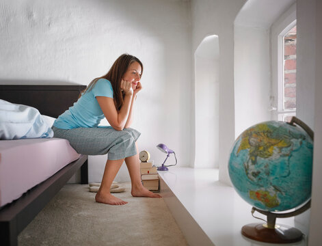 Woman in pajamas sits on bed, gazing thoughtfully at a globe by a window. Germany